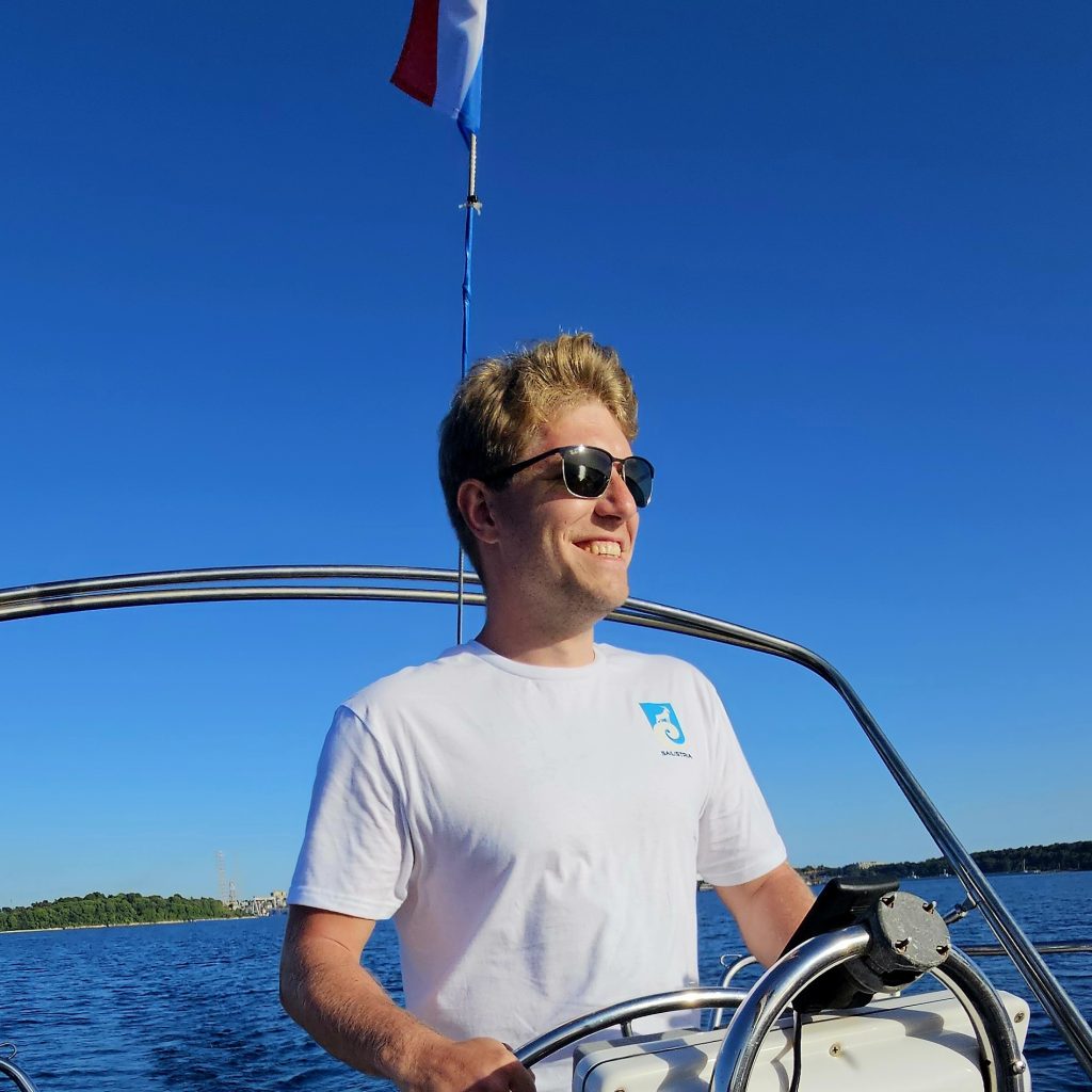 Andrej, co-owner of Sail Istria, standing on a sailboat, enjoying a sunny day on the water with a blue sky in the background.