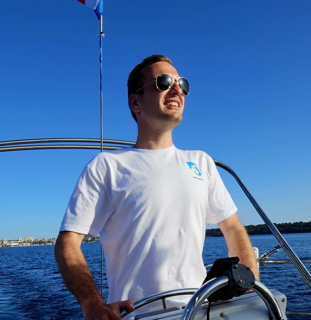 Anton, co-owner of Sail Istria, standing at the helm of a sailboat, smiling under a clear blue sky with the sea in the background.