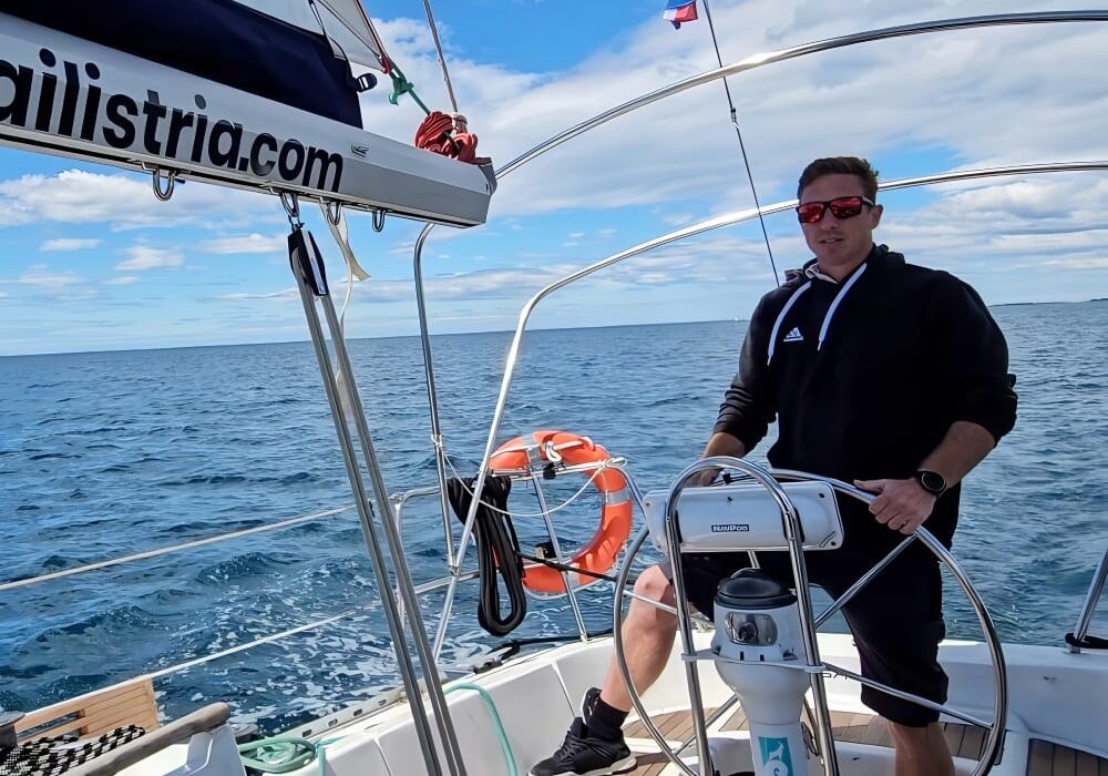 Profesionalni kapetan tvrtke Sail Istria upravlja jedrilicom pod plavim nebom, s vidljivom hrvatskom zastavom.
